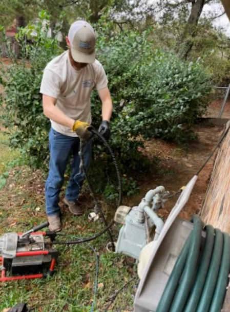 Worker operating a power tool beside coiled hose and equipment in a garden area