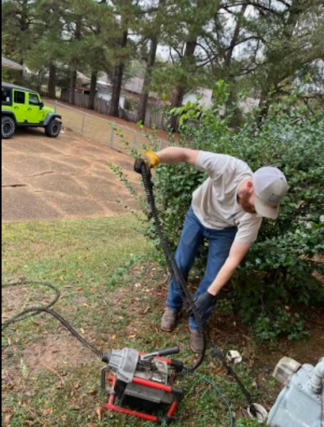 Person using a chainsaw to trim a hedge beside a driveway, with a green vehicle in the background.