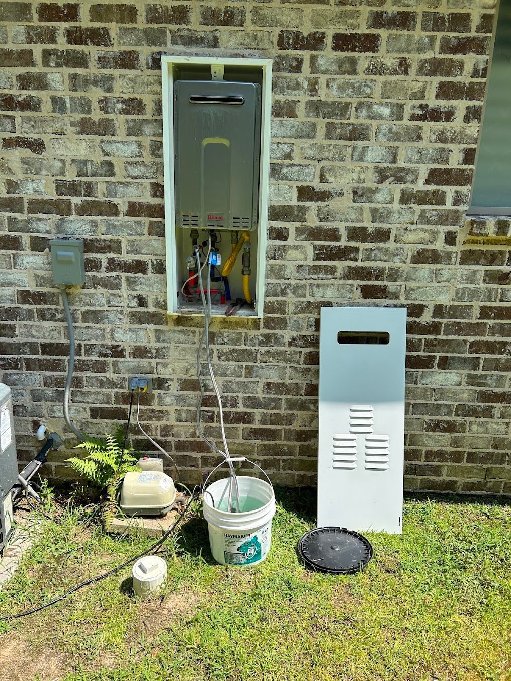 Outdoor utility meters and equipment mounted on a brick wall with a bucket and grass below