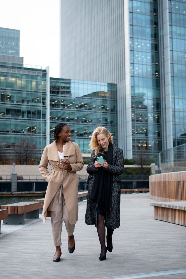 Two women are walking down a sidewalk in front of tall buildings.