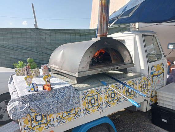 Un forno a legna per pizza all'aperto, posizionato su un tavolo decorato in un mercato di strada, con le fiamme visibili all'interno.