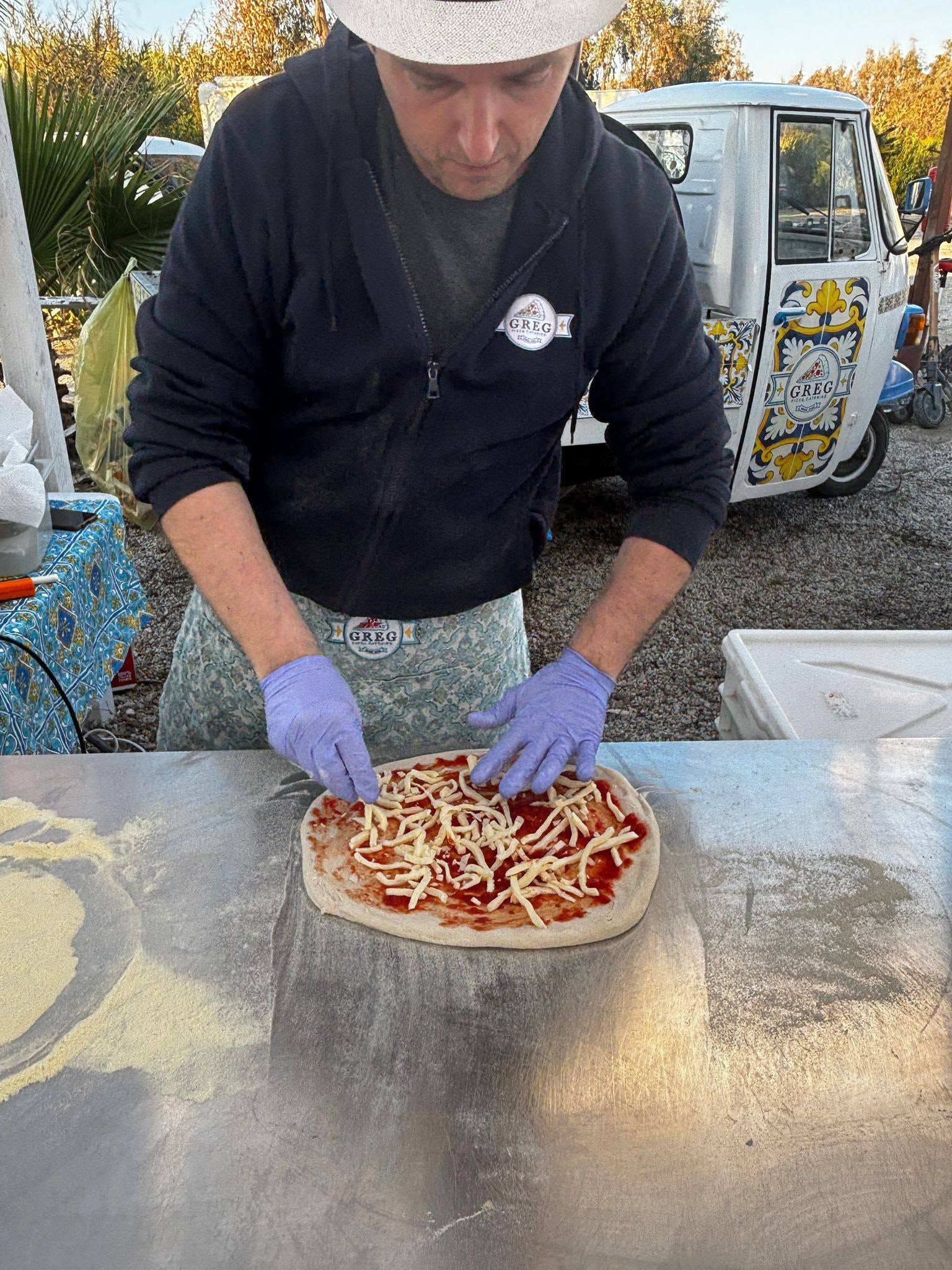 Un operaio con i guanti viola spalma il formaggio su una pizza su un tavolo di preparazione all'aperto.