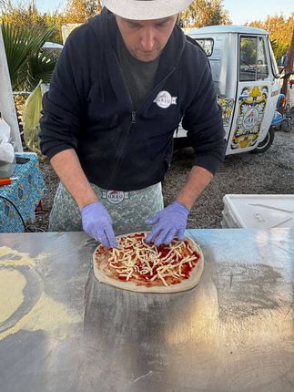 Una persona con un elmetto e guanti viola spalma gli ingredienti su una pizza all'aperto presso un food truck.