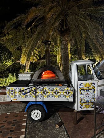 Un forno per pizza decorativo su un piccolo camioncino, illuminato di notte sotto le palme e rivestito di piastrelle con motivi blu-gialli.