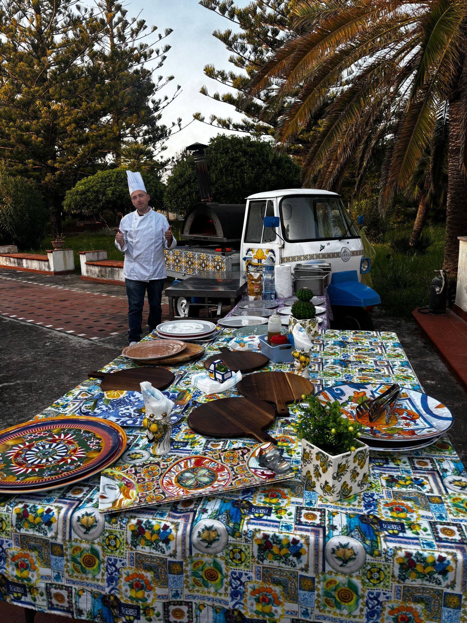 Un tavolo da buffet all'aperto con tovaglie colorate, piatti e uno chef in piedi accanto a un camioncino bianco in un giardino.