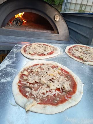 Le pizze, farcite con formaggio e salsa, sono disposte davanti a un forno a legna con fiamme visibili all'interno.