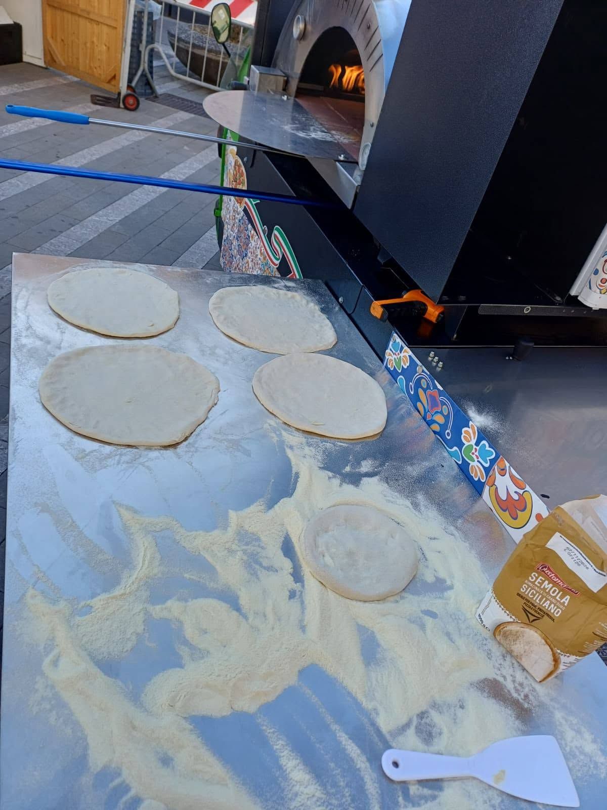 Quattro dischi di pasta rotondi su un tavolo infarinato accanto a un forno a legna con gli ingredienti per la pizza.