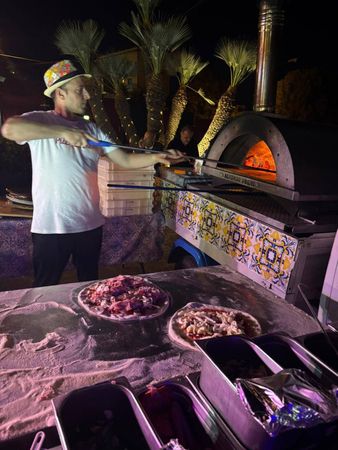 Una persona si occupa di un forno a mattoni incandescente con teglie di cibo preparato in una postazione di cottura notturna all'aperto.