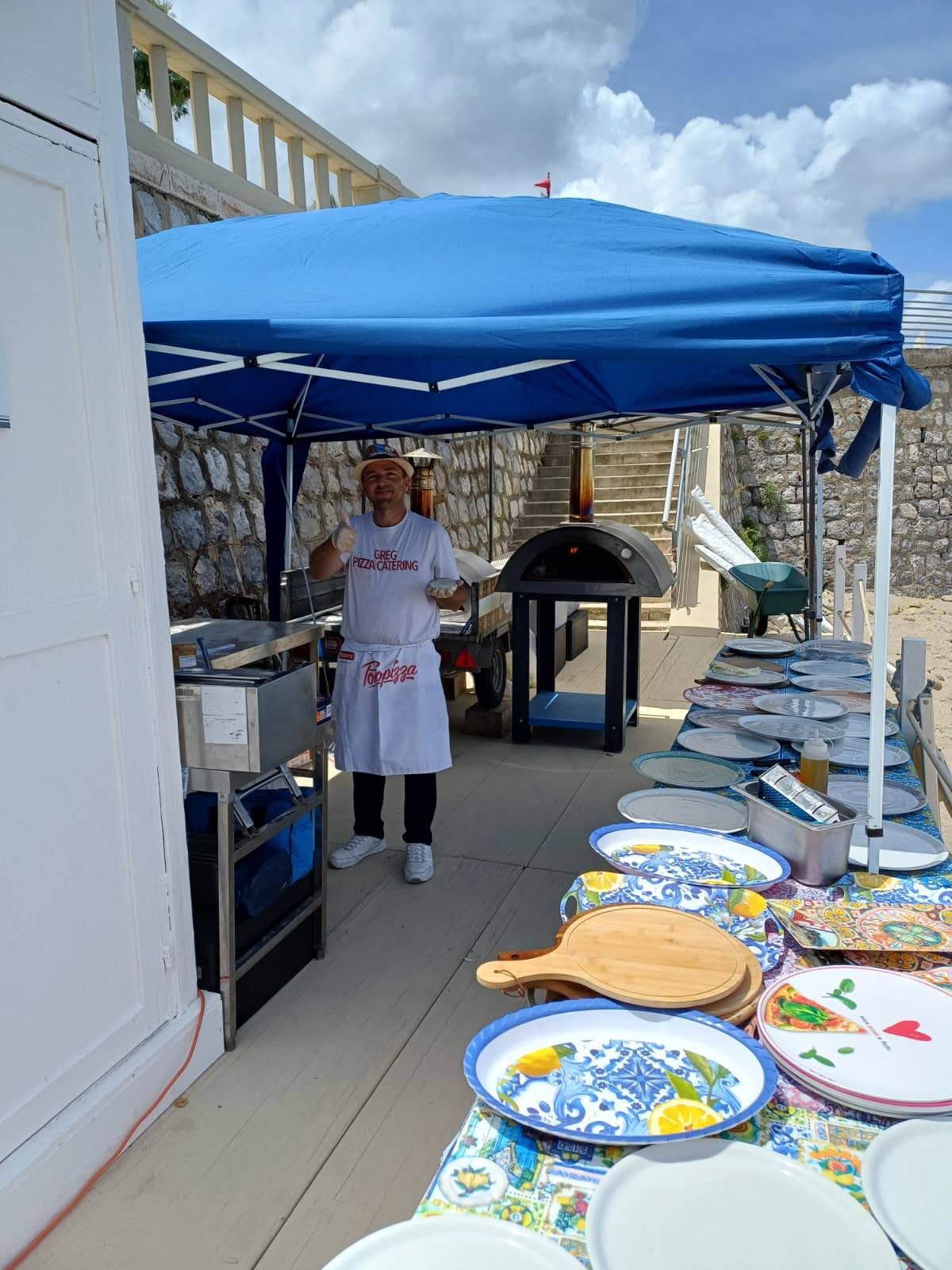 Buffet all'aperto sotto una tettoia blu, con una persona in grembiule bianco in piedi accanto ai tavoli con le pietanze.
