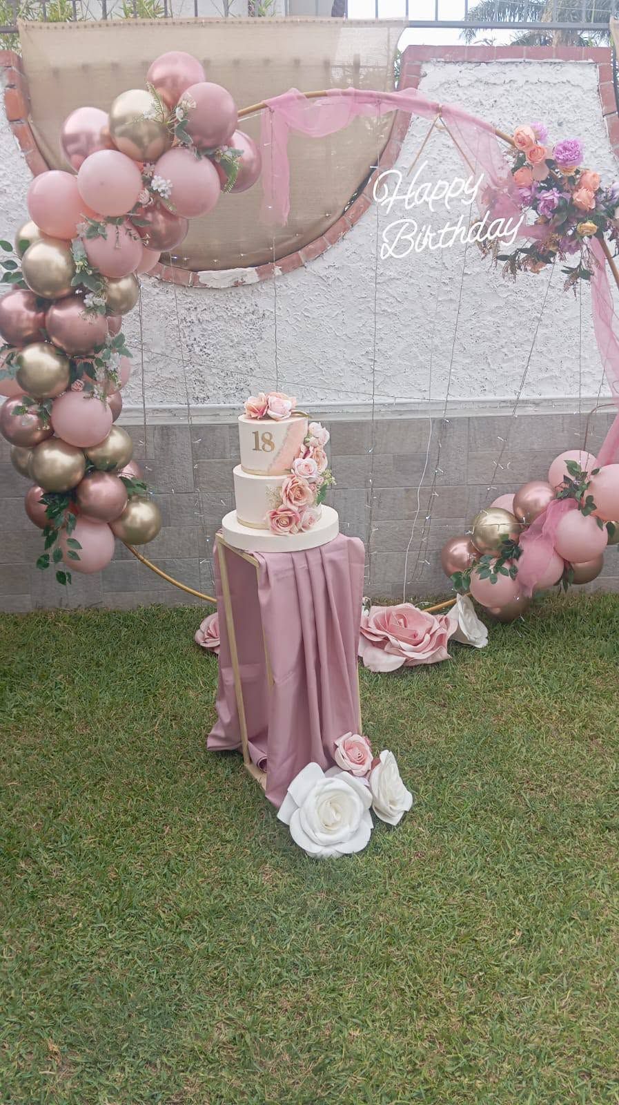 Composizione floreale con palloncini rosa e torta su un supporto drappeggiato, in un'ambientazione da giardino.