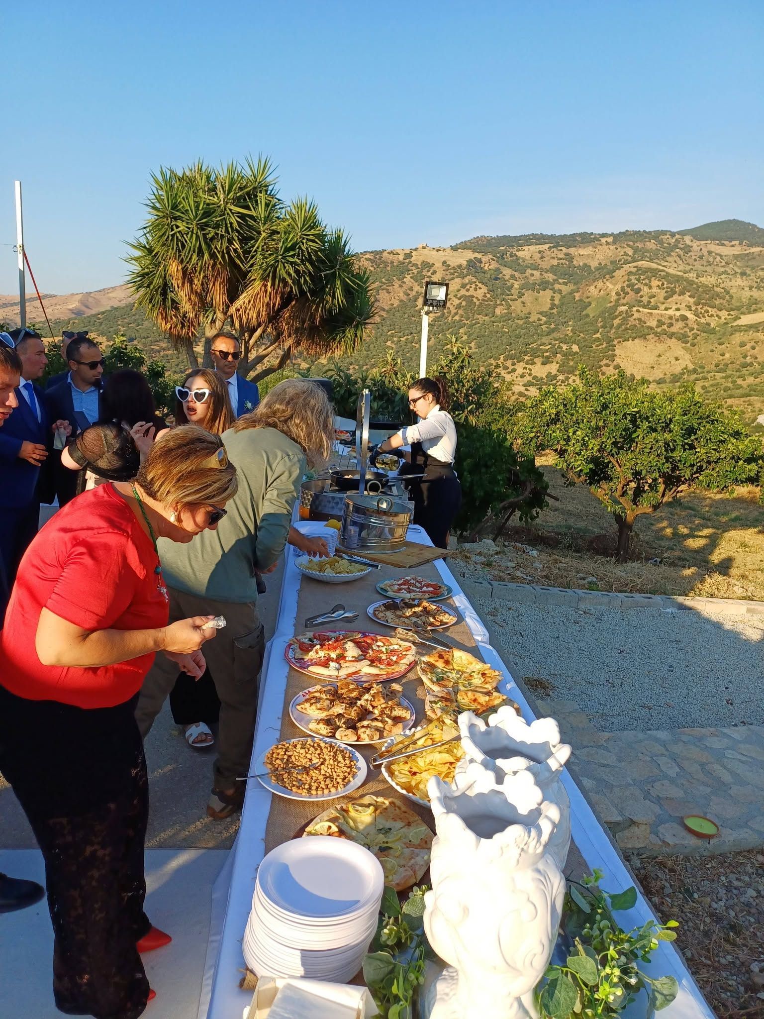 Tavolo da buffet all'aperto con persone che servono cibo, sullo sfondo di uno splendido panorama collinare.