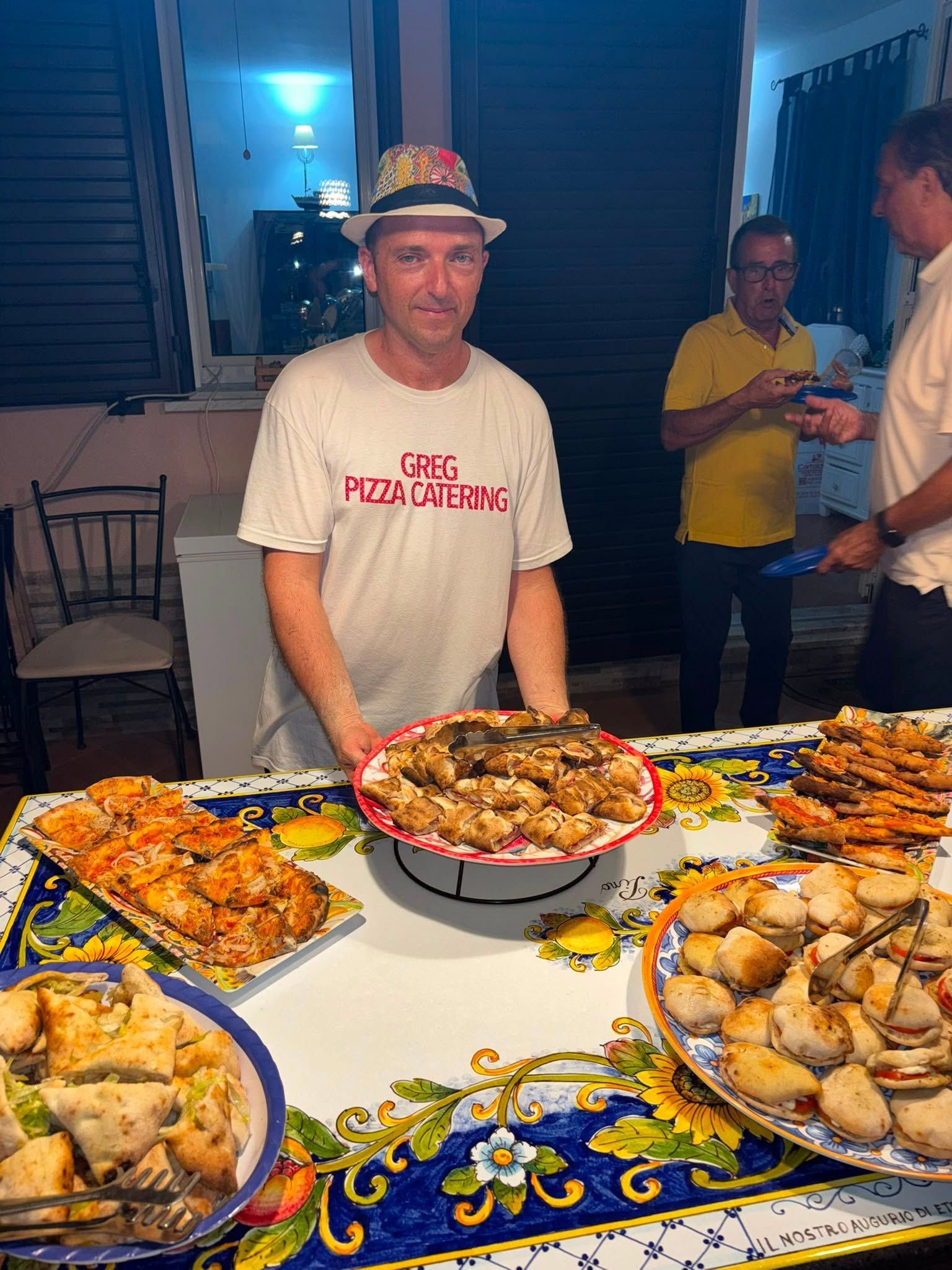 Un uomo in piedi dietro un tavolo da buffet con vassoi di cibo e una tovaglia decorativa durante un evento al chiuso.