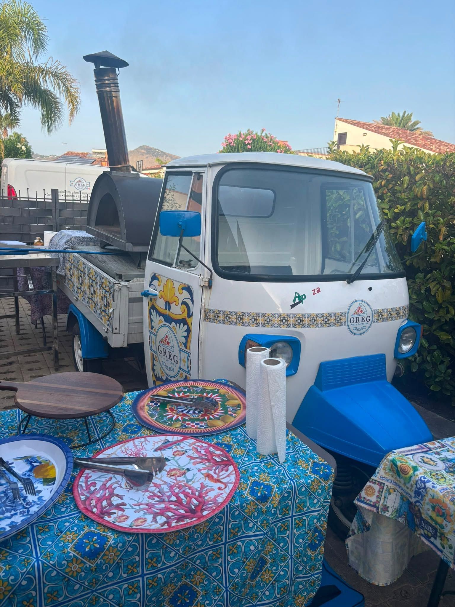 Un piccolo camioncino bianco presso una bancarella di un mercato all'aperto con tovaglie e pentole colorate, sotto un cielo limpido.