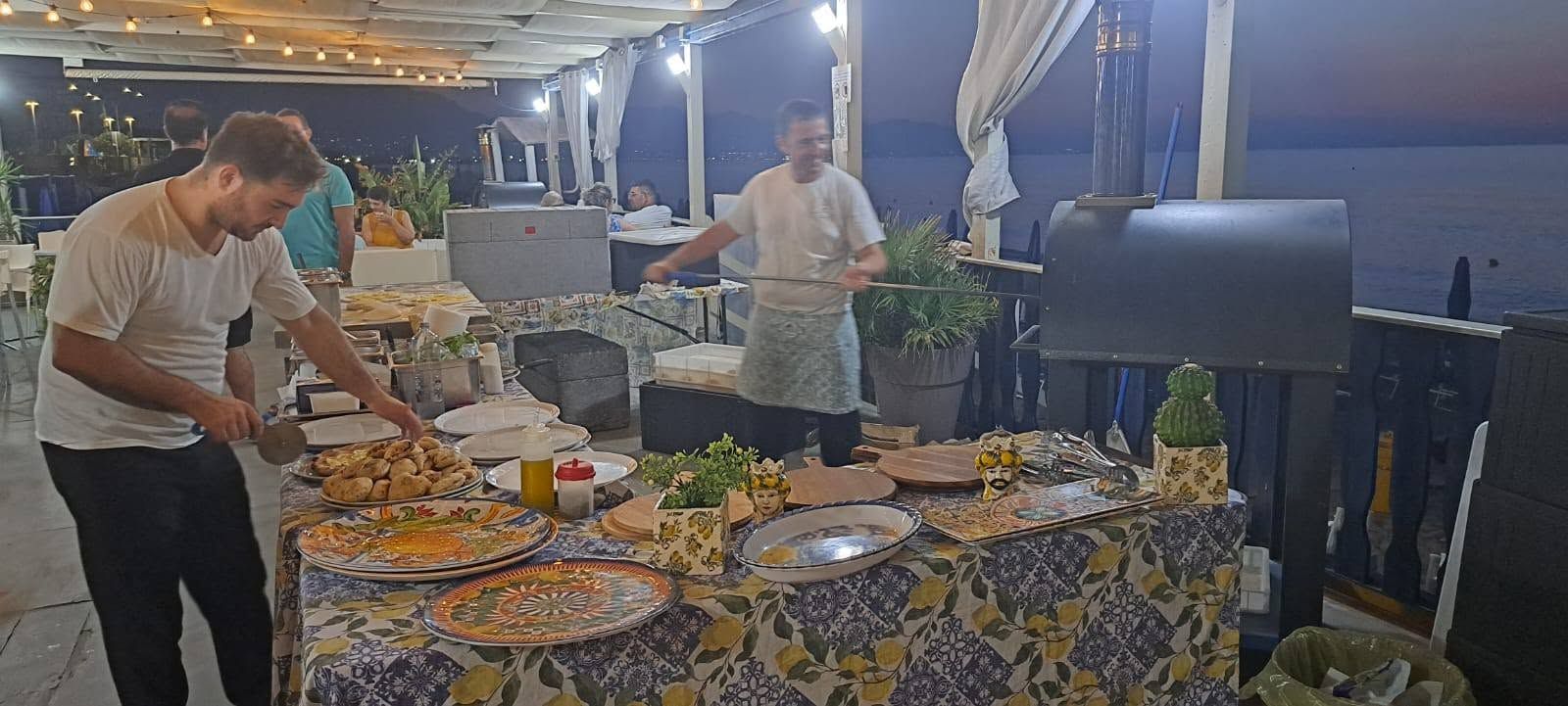 Due uomini servono cibo a un buffet illuminato da lucine su una terrazza all'aperto al tramonto.