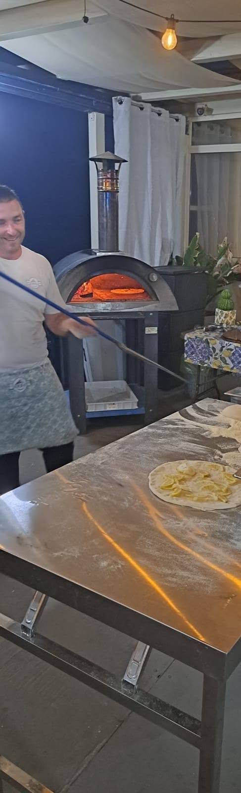 Una persona tiene in mano una pala per pizza con una pizza appena sfornata in un forno a legna, accanto a un tavolo di lavoro ricoperto di impasto.