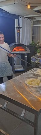 Una persona tiene in mano una pala per pizza con una pizza appena sfornata in un forno a legna, accanto a un tavolo di lavoro ricoperto di impasto.