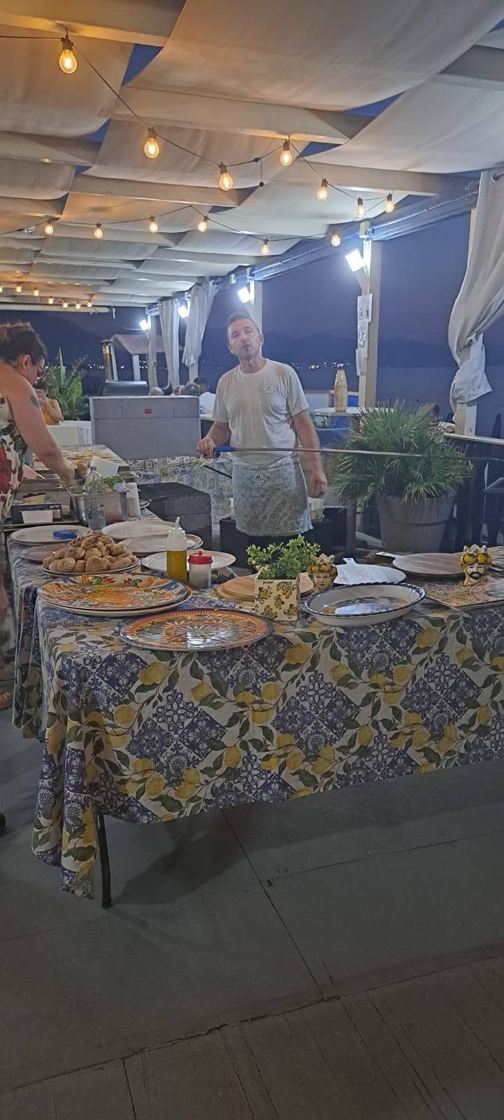 Un tavolo da buffet sotto un baldacchino con luci decorative, ricoperto di cibo e piante, con una persona in piedi dietro di esso.
