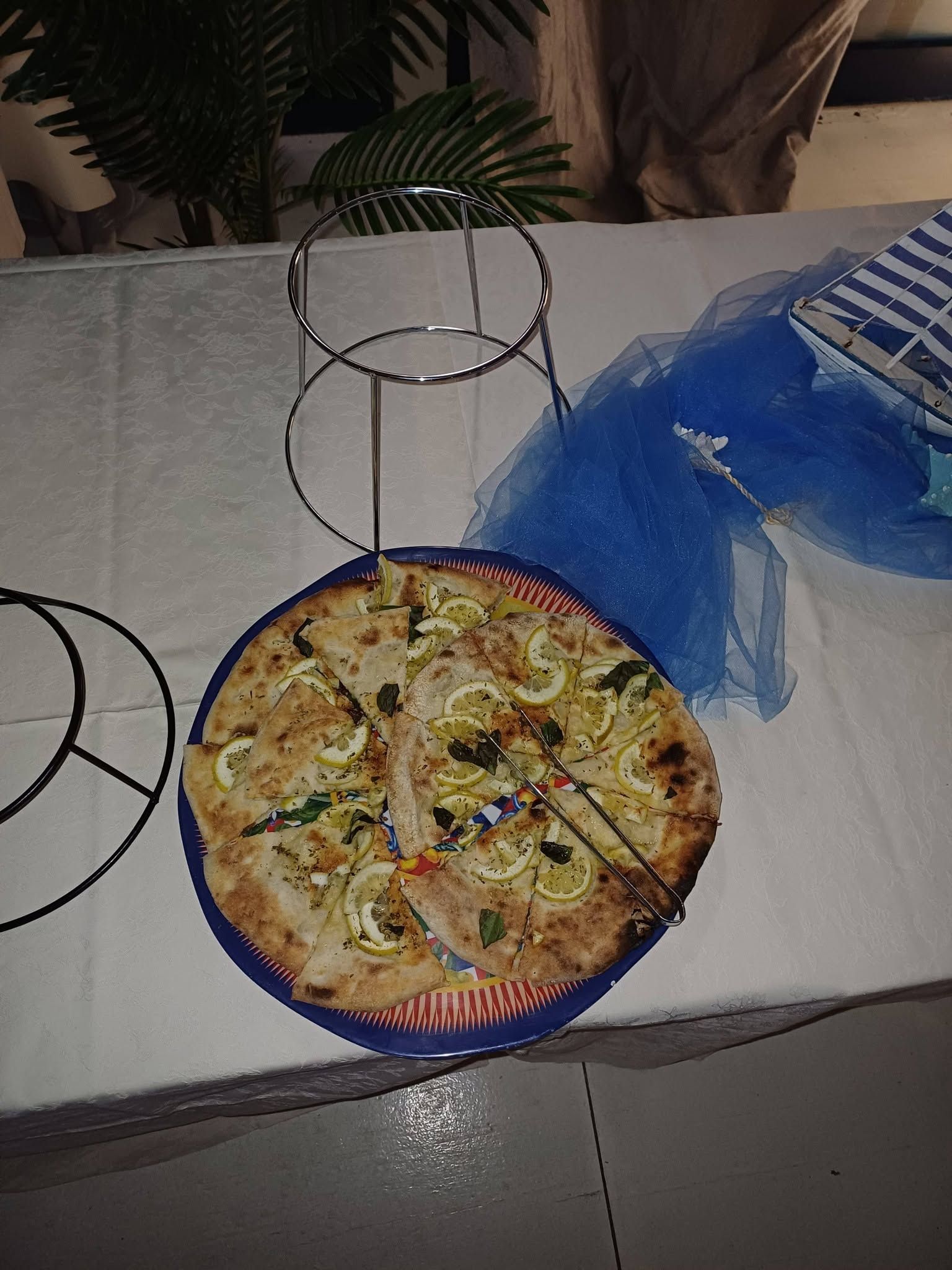 Una pizza su un tavolo accanto a una borsa blu e alla struttura di una sedia in filo metallico nero in un ambiente interno informale.