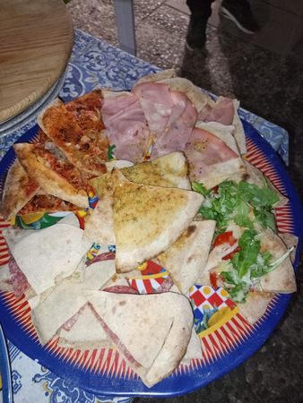Un assortimento di piadine e panini su un piatto colorato, con prosciutto, verdure e formaggio su una tovaglia decorata.