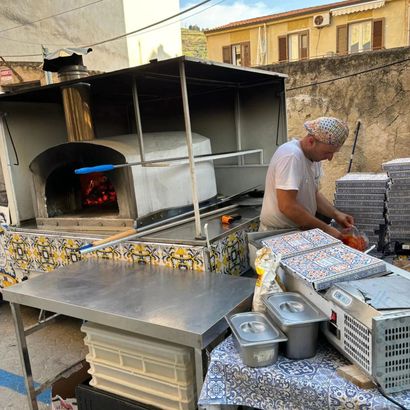 Un uomo prepara dei vassoi accanto a un forno a legna presso una bancarella di cibo all'aperto.