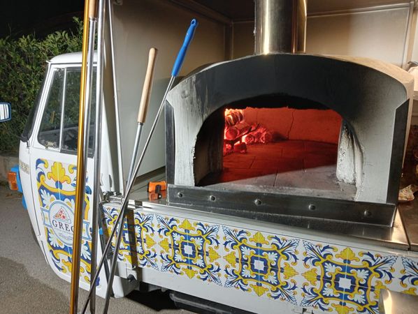 Forno per pizza a legna in una cucina piastrellata allestita su un camion, con utensili e fuoco scoppiettante all'interno.