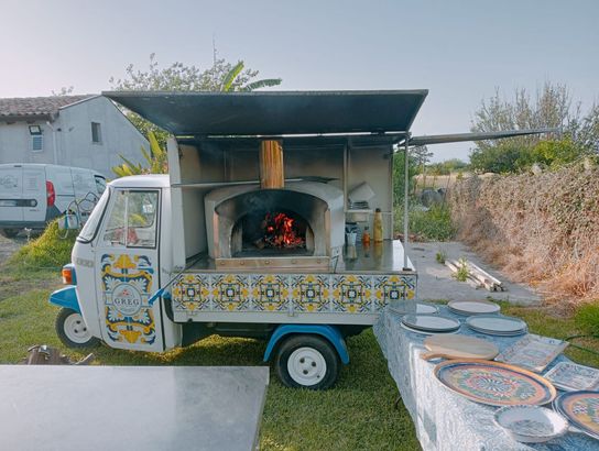 Un camioncino decorato con forno a legna per pizza, parcheggiato all'aperto, con pizze in cottura all'interno.