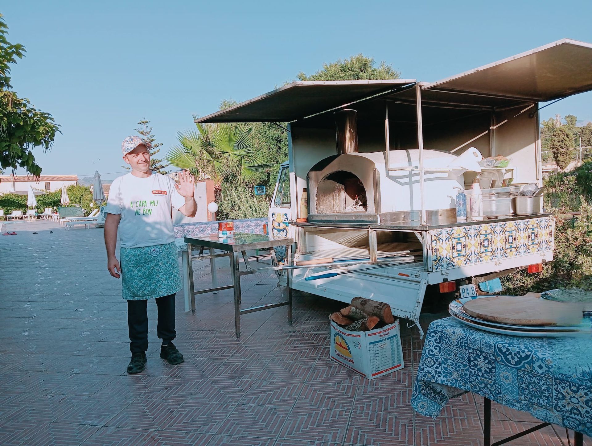 Bancarella all'aperto con una persona in piedi accanto a un forno a legna mobile e tavoli sotto delle tettoie.