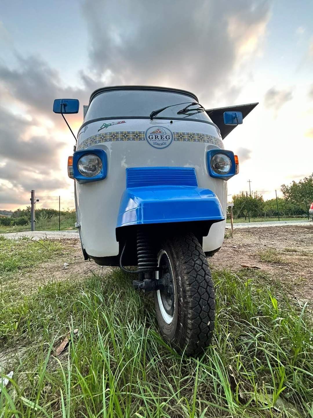 Tuk-tuk blu e bianco parcheggiato sull'erba sotto un cielo nuvoloso, vista frontale