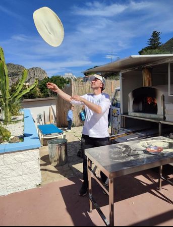Una persona lancia l'impasto della pizza accanto a un forno a legna all'aperto su un patio soleggiato.
