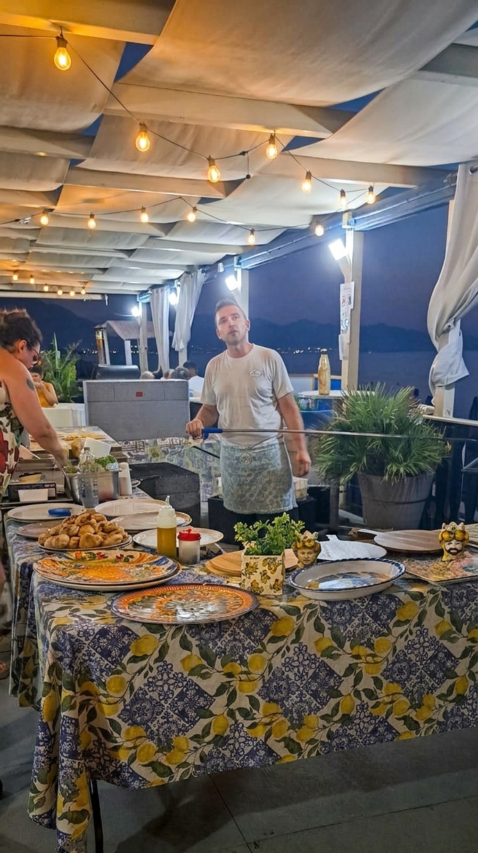 Tavolo da buffet sotto una tettoia esterna illuminata, con piatti, vassoi e due persone che preparano il cibo.
