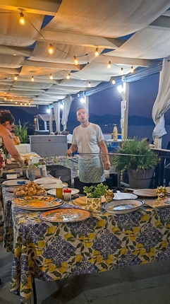 Tavolo da buffet sotto una tettoia esterna illuminata, con piatti, vassoi e due persone che preparano il cibo.