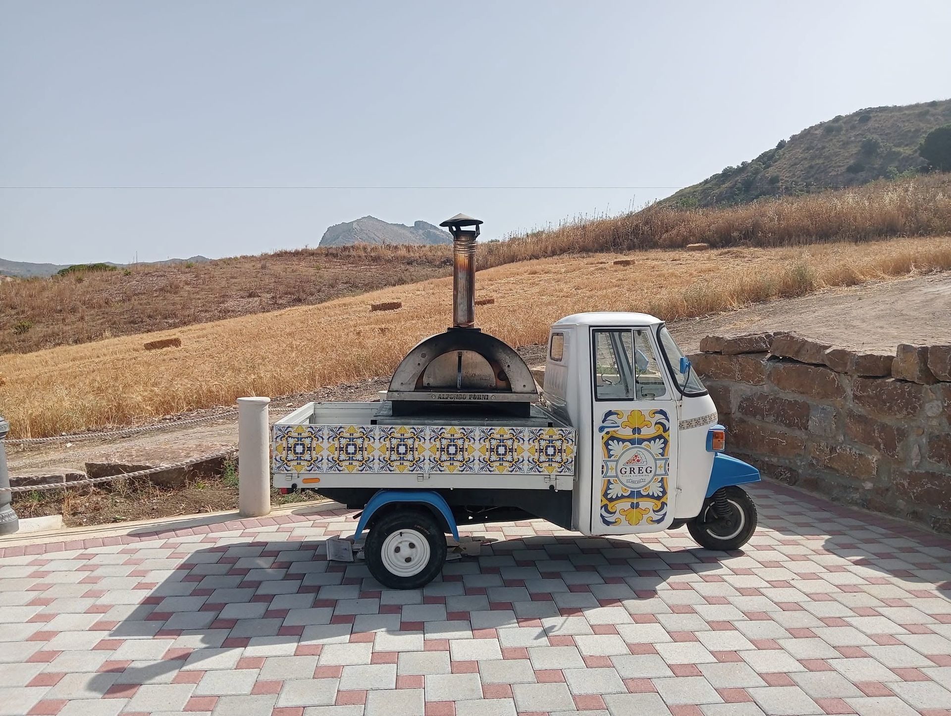 Un piccolo furgone bianco adibito a uso commerciale, con forno a legna, è parcheggiato su un patio piastrellato in una zona collinare arida.