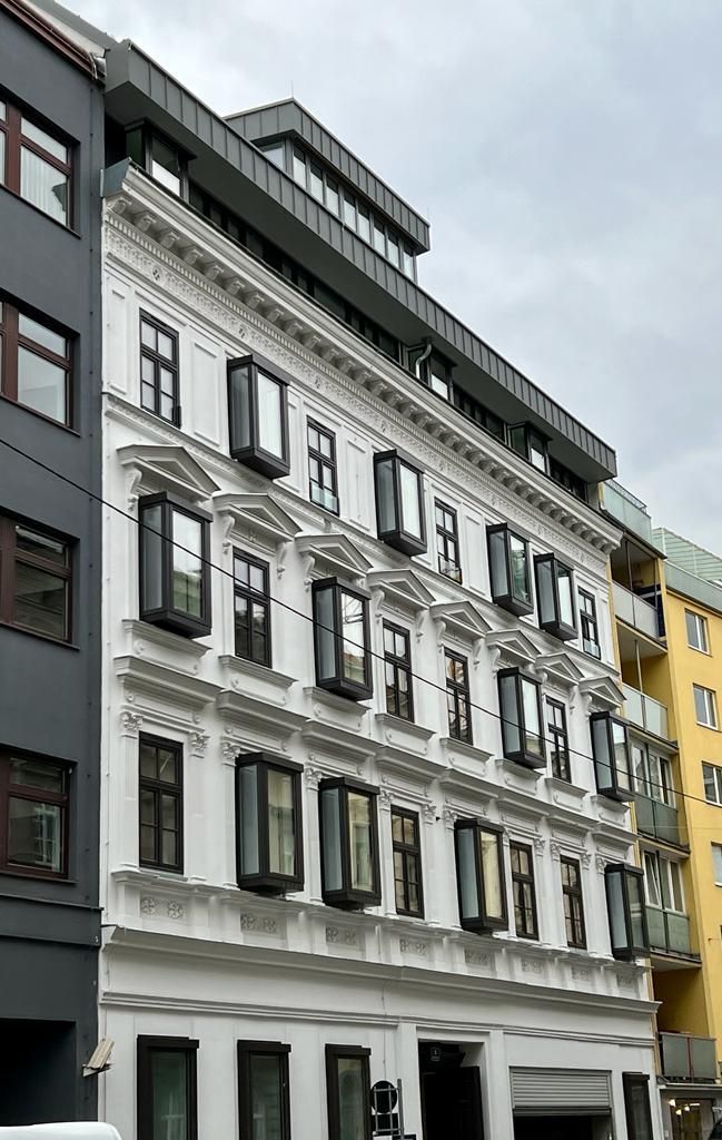 Weißes Gebäude mit dunklen Fensterrahmen und einem modernen grauen Dachaufsatz.