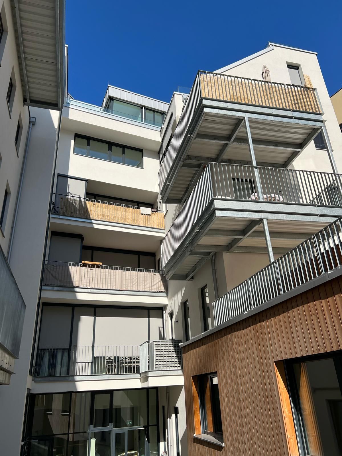 Modernes Mehrfamilienhaus mit Balkonen, grau-weiße Fassade, sonniger Tag.