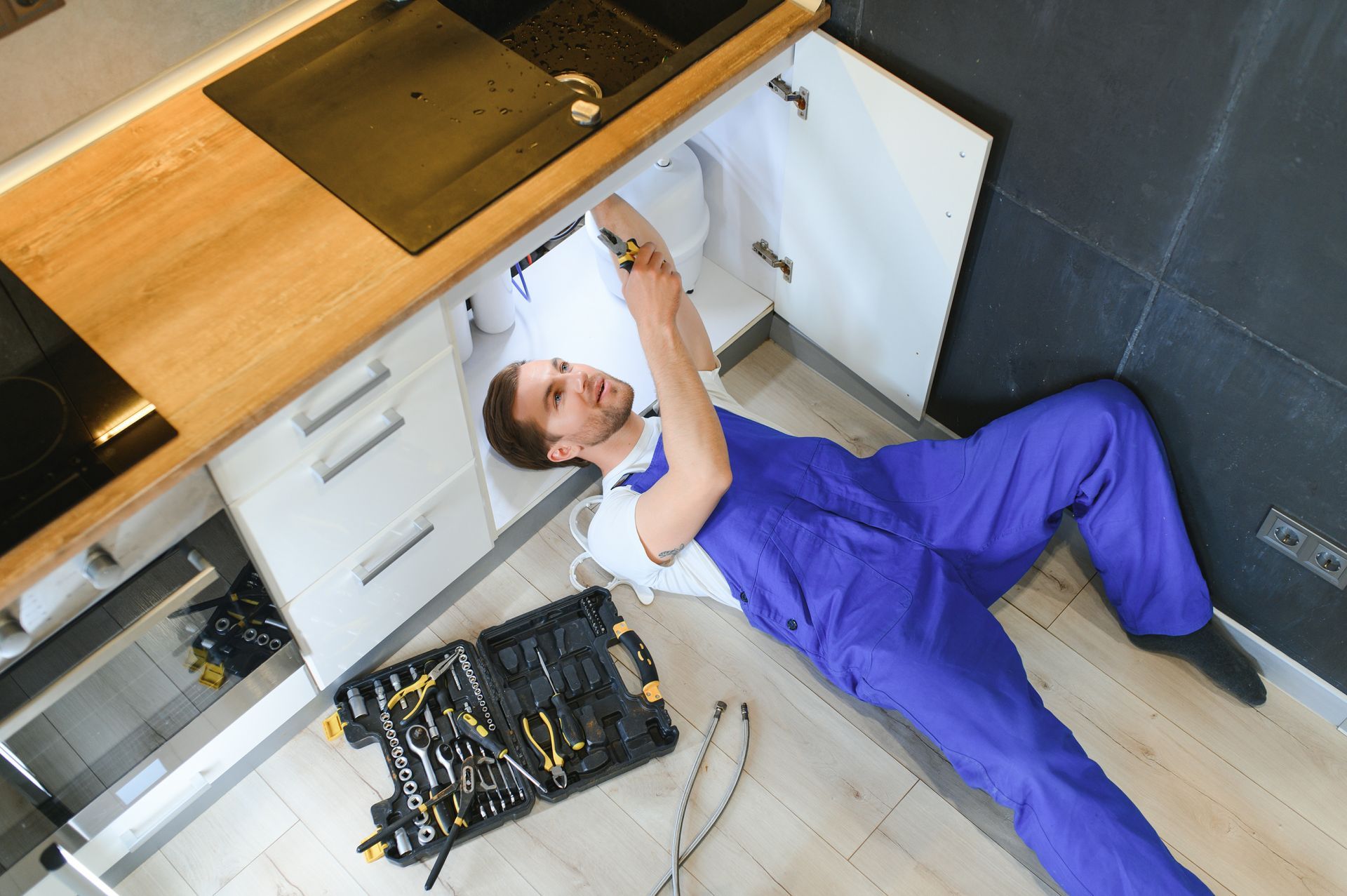 A person in blue overalls repairs something under a kitchen sink, with tools nearby.