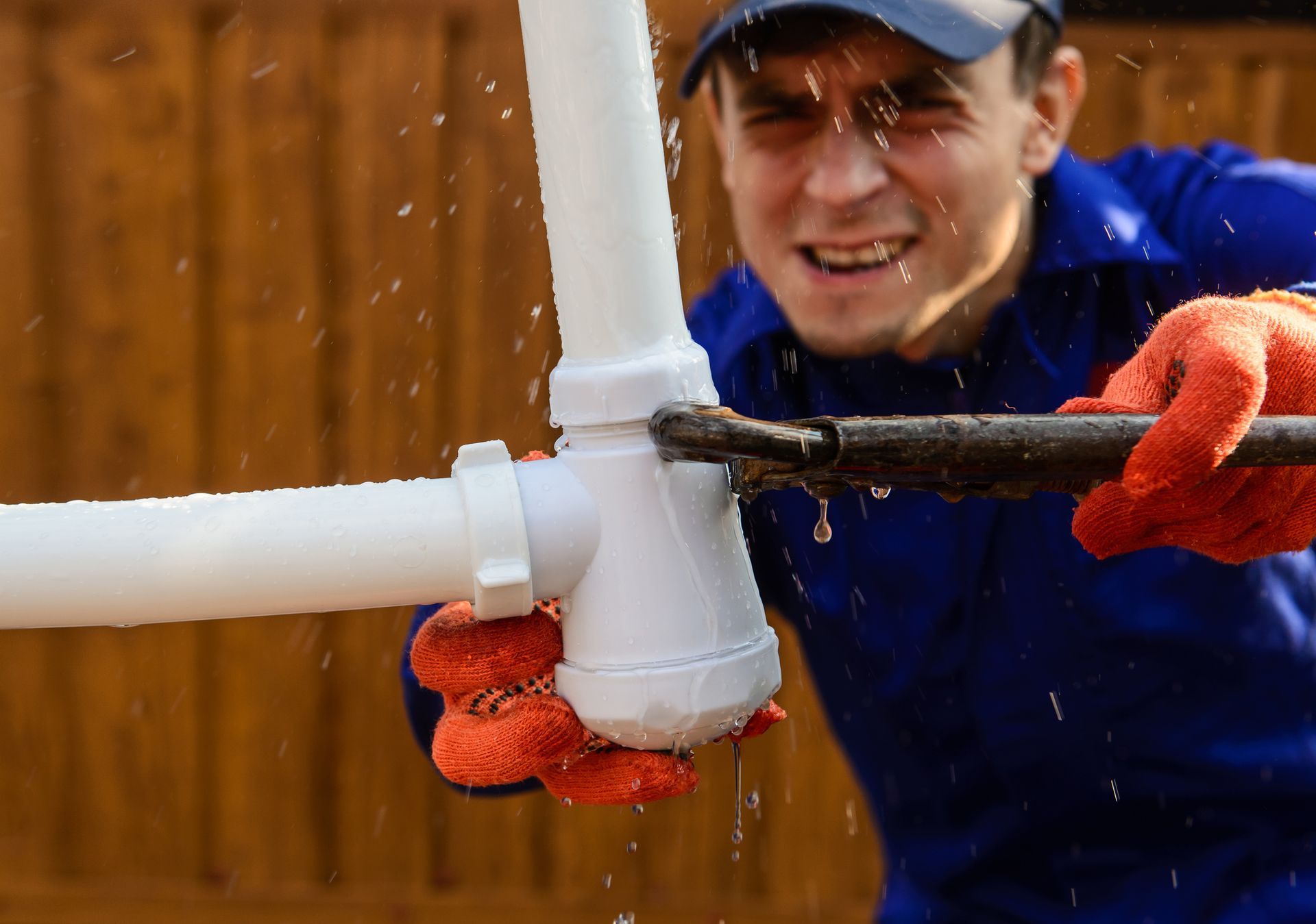 Plumber struggles to fix a leaky pipe, water spraying. He wears gloves and a blue uniform.