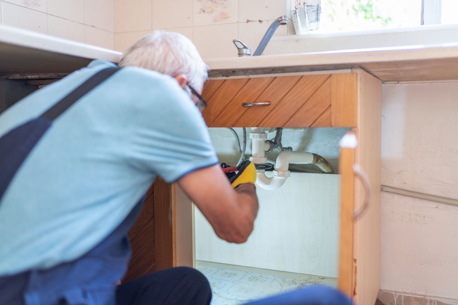 Plumber in blue workwear repairs pipes under a kitchen sink.