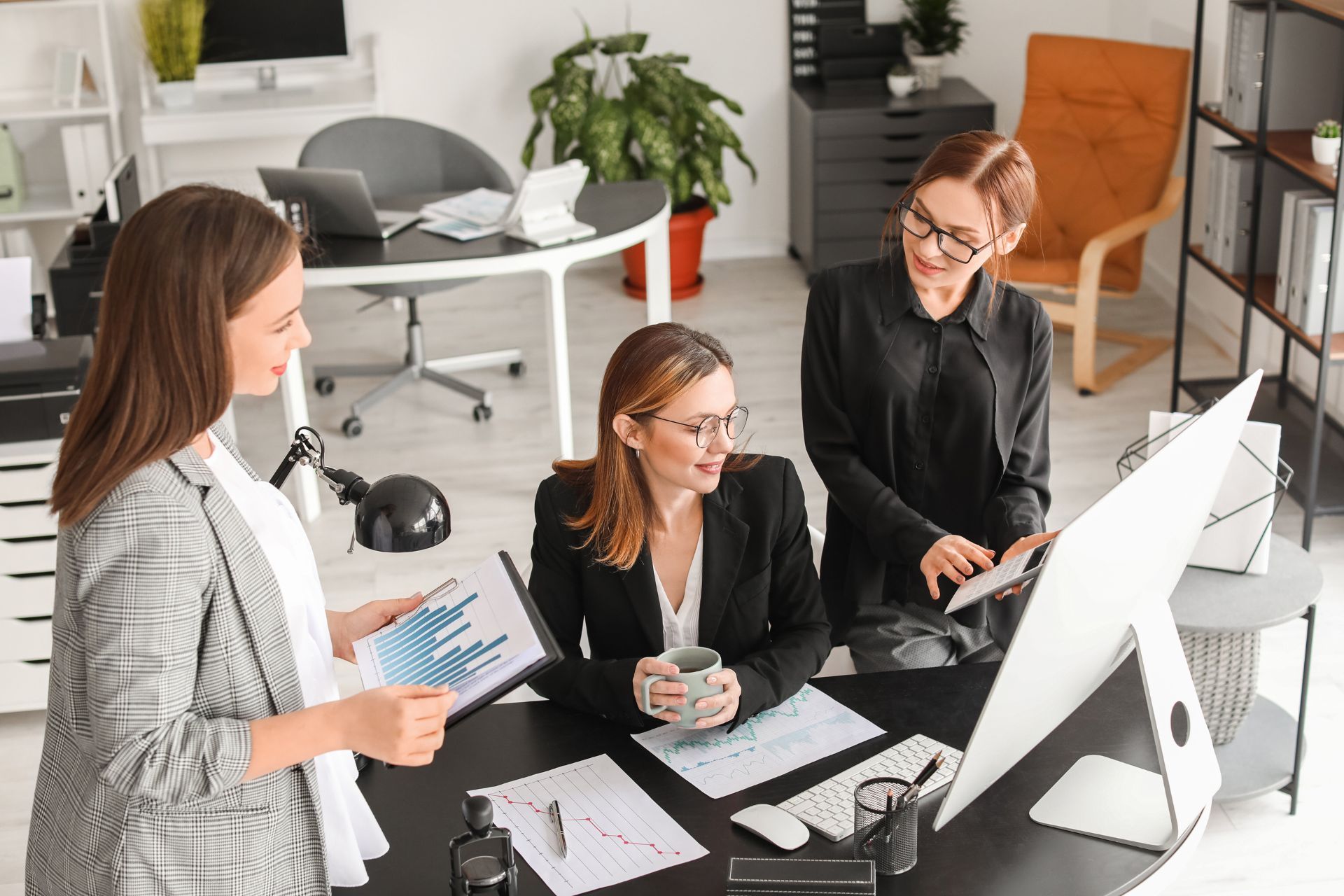 three women are sitting at a desk in an office looking at a computer screen .