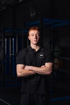 Man standing with arms crossed in a dark gym, wearing a black polo shirt, facing the camera.