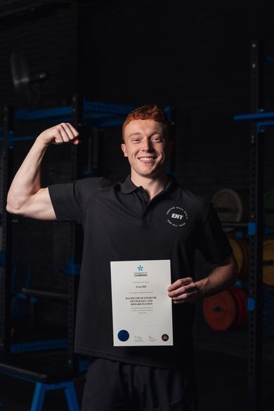 Smiling man in black polo flexes beside a certificate in a dim gym