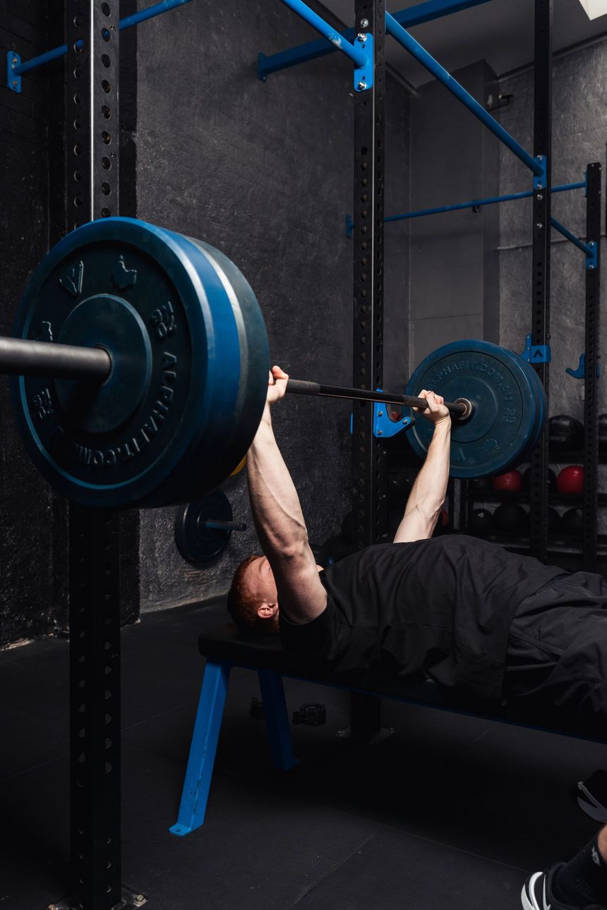 Person bench pressing a barbell in a gym, wearing black, with blue weight plates.