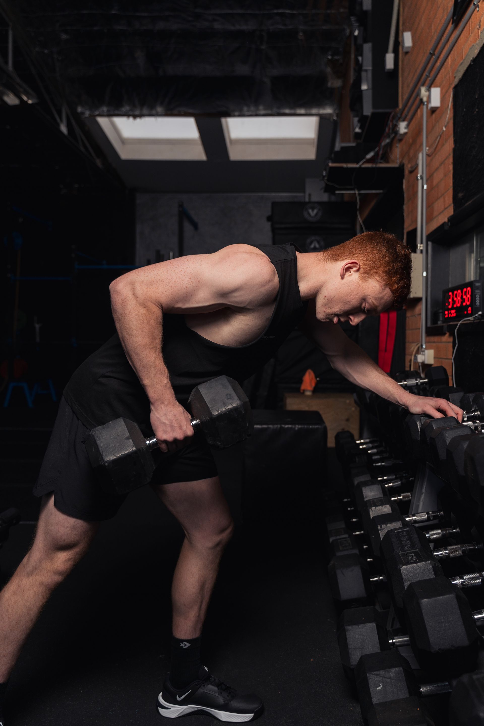 Man lifting a dumbbell in a dark gym, bent over beside a rack of weights.