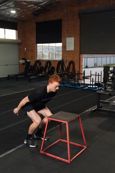 Person jumping over a red plyometric box in a gym.