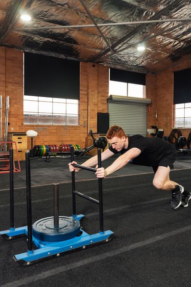 Person pushing a blue weighted sled across a gym floor indoors
