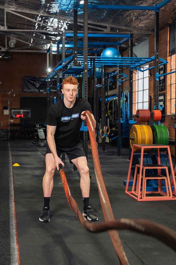 Person exercising with battle ropes in a gym, with colorful equipment and metal rigging in the background.