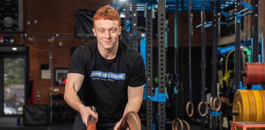Person in a gym, smiling while using battle ropes, with weights and equipment in the background.