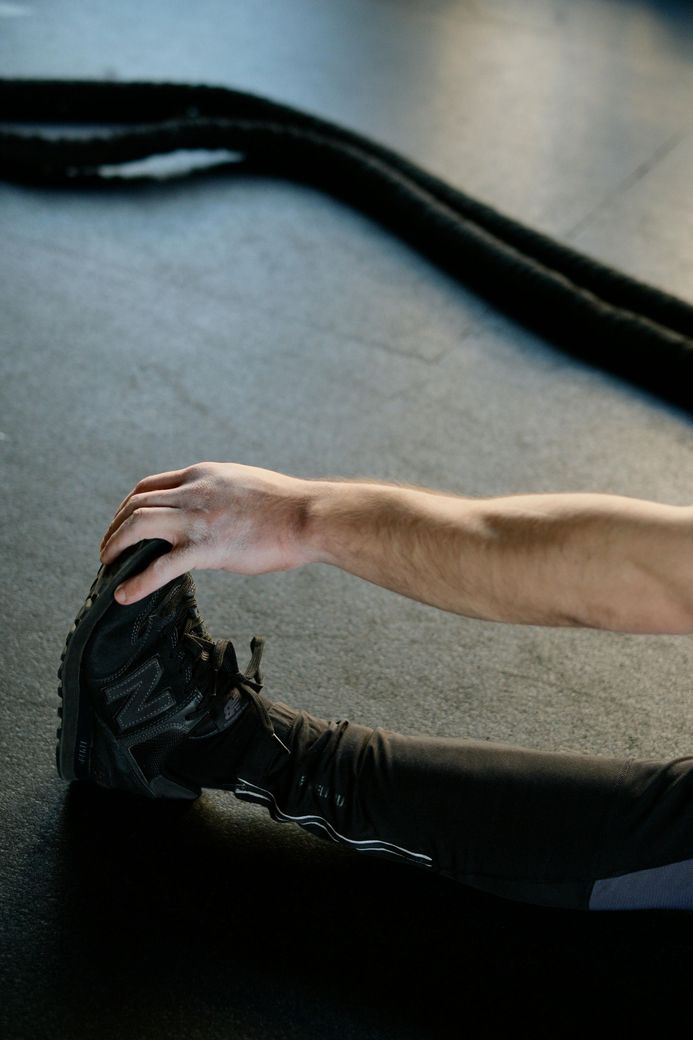 Hand reaching toward a black shoe on a gray floor, with a black strap in the background.