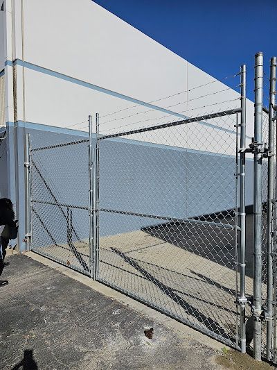 Chain-link gate with barbed wire on a concrete lot beside a white building under a blue sky