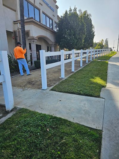 Worker in orange shirt painting a white fence along a sidewalk beside a building and lawn