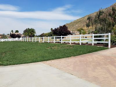 White fence along a grassy path beside a paved road, with hills and a blue sky in the background.
